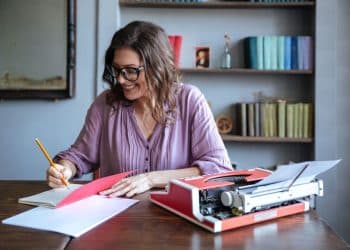 Mulher escrevendo anotações ao lado de uma máquina de escrever em um ambiente com livros, representando carreiras para quem gosta de escrever.