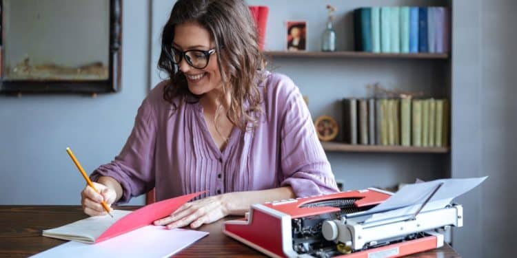 Mulher escrevendo anotações ao lado de uma máquina de escrever em um ambiente com livros, representando carreiras para quem gosta de escrever.