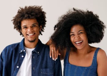 Dois adultos sorrindo, um com camisa jeans e outro com vestido jeans, fundo claro.