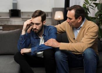 Pai consola filho adulto em conversa emocional no sofá de casa.