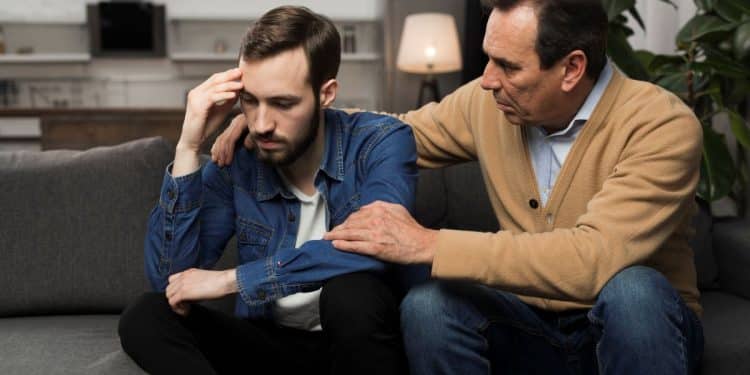 Pai consola filho adulto em conversa emocional no sofá de casa.