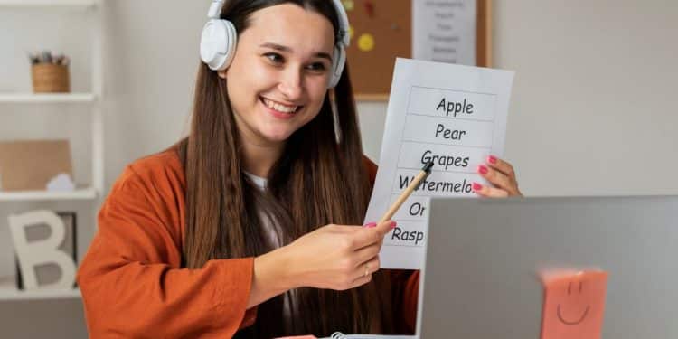 Jovem professora usando fones e sorrindo enquanto aponta para lista de frutas em inglês durante aula online.