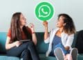 Amigas em sofá seguram placa com logo do WhatsApp sorrindo.