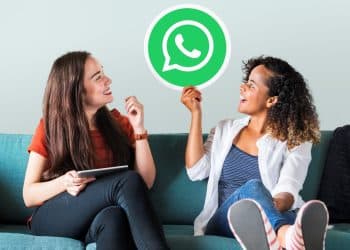 Amigas em sofá seguram placa com logo do WhatsApp sorrindo.