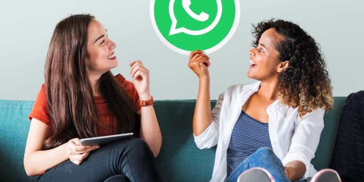 Amigas em sofá seguram placa com logo do WhatsApp sorrindo.