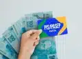 Mão segurando cartão do PIS Pasep da Caixa em frente a cédulas de R$ 100, simbolizando o pagamento do abono salarial em 2026