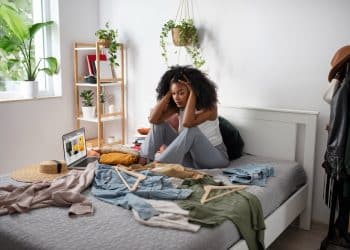 Mulher sentada na cama com as mãos na cabeça, cercada de roupas, cabides e objetos espalhados, representando a frustração diante de um ambiente desorganizado