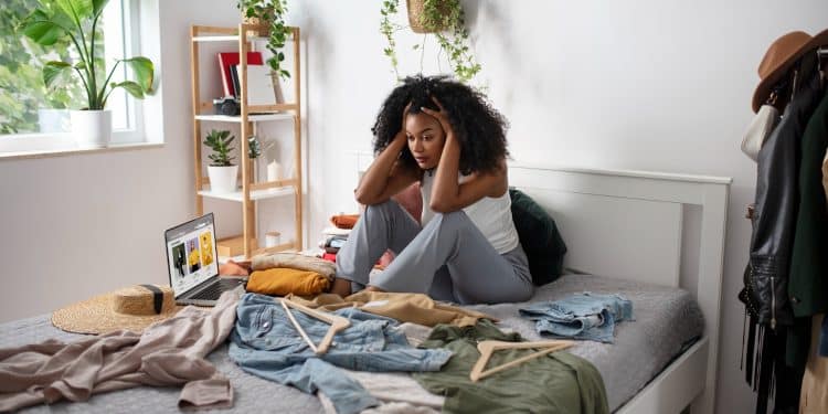 Mulher sentada na cama com as mãos na cabeça, cercada de roupas, cabides e objetos espalhados, representando a frustração diante de um ambiente desorganizado