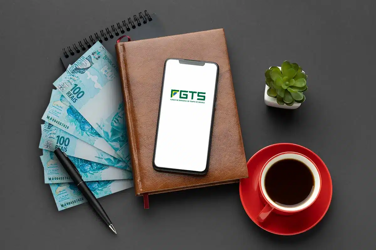 Imagem ilustrativa mostrando o aplicativo FGTS aberto em um celular, com cédulas de R$100, uma caneta, uma planta e uma xícara de café, representando a facilidade de solicitar e receber o Saque-Aniversário.