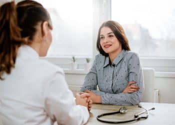 Assistente social em atendimento presencial com uma mulher em um consultório iluminado, representando a atuação profissional na área da saúde.