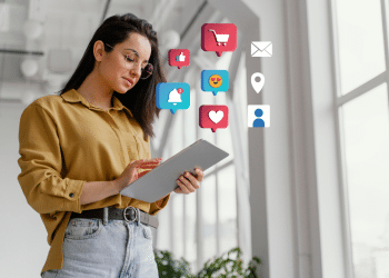 Jovem mulher de camisa mostarda e óculos segura tablet cinza com ícones de like, coração, notificação e e-mail ao redor