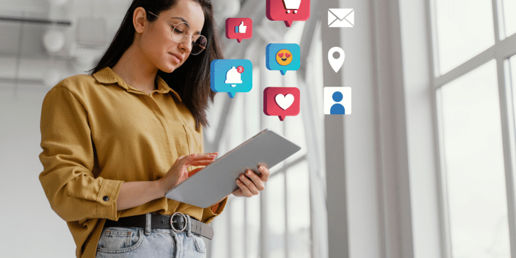 Jovem mulher de camisa mostarda e óculos segura tablet cinza com ícones de like, coração, notificação e e-mail ao redor