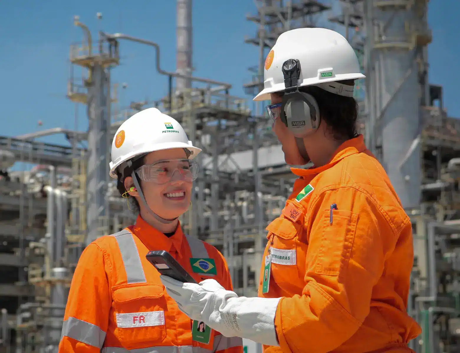 Funcionárias da Petrobras conversando em ambiente industrial, com uniforme de segurança laranja e equipamentos de proteção em uma planta de processamento.