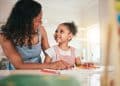 Mãe auxiliando a filha pequena em atividade escolar enquanto conversam e demonstram conexão emocional em casa.