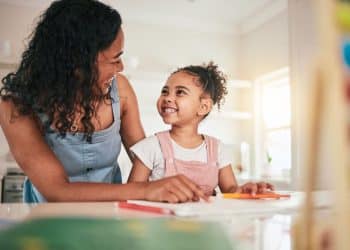 Mãe auxiliando a filha pequena em atividade escolar enquanto conversam e demonstram conexão emocional em casa.