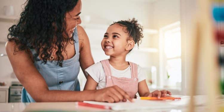 Mãe auxiliando a filha pequena em atividade escolar enquanto conversam e demonstram conexão emocional em casa.