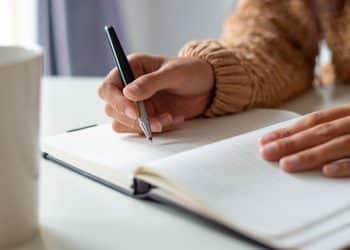 Close focado nas mãos de uma pessoa de pele morena claro, usando um blusão de lã marrom claro. A mão direita segura uma caneta esferográfica de metal prata e preto e escreve na página de um caderno de anotações com capa escura, aberto sobre uma mesa branca. A mão esquerda está apoiada sobre o caderno. À esquerda, uma caneca branca está desfocada. O fundo é neutro e suave.