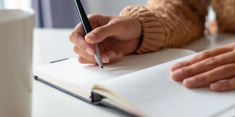 Close focado nas mãos de uma pessoa de pele morena claro, usando um blusão de lã marrom claro. A mão direita segura uma caneta esferográfica de metal prata e preto e escreve na página de um caderno de anotações com capa escura, aberto sobre uma mesa branca. A mão esquerda está apoiada sobre o caderno. À esquerda, uma caneca branca está desfocada. O fundo é neutro e suave.