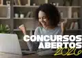 Mulher negra de cabelos cacheados sorri e comemora com os braços levantados em frente a um notebook aberto, com os textos "Concursos Abertos 2026" sobrepostos em branco e amarelo, representando candidatos que podem se inscrever no concurso público da Prefeitura Municipal de Guzolândia — SP, com vagas para Escriturário, Nutricionista Clínico, Médico Pediatra e outros cargos em 2026