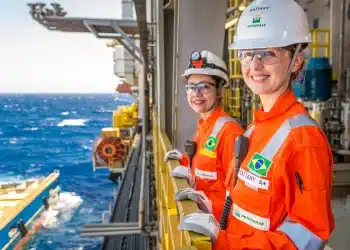 Funcionárias da Petrobras sorrindo em plataforma offshore, com uniforme de segurança laranja, em ambiente marítimo.