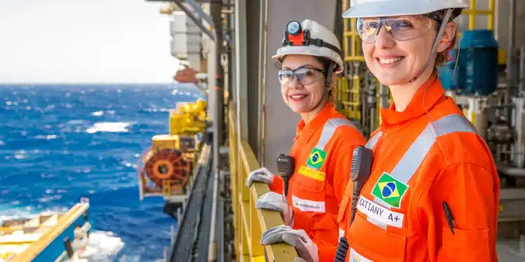 Funcionárias da Petrobras sorrindo em plataforma offshore, com uniforme de segurança laranja, em ambiente marítimo.