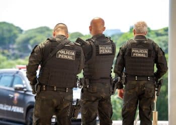 Três policiais penais de costas usando coletes pretos com inscrição "Polícia Penal" e viatura estacionada