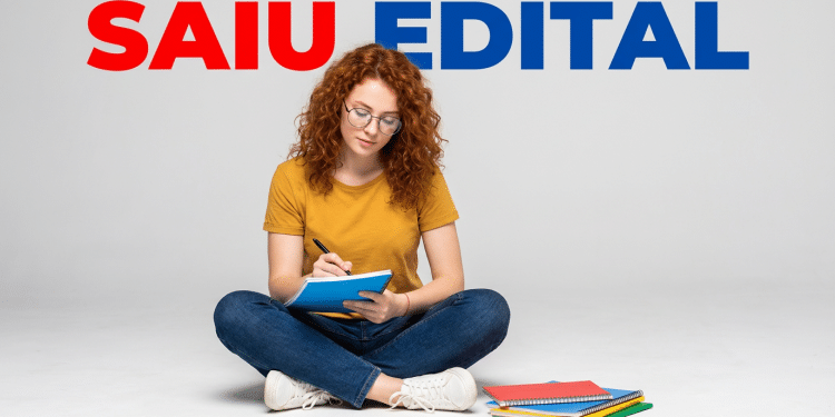 Jovem ruiva de óculos sentada no chão, escrevendo em caderno azul, com pilha de cadernos coloridos ao lado e texto "Saiu Edital"