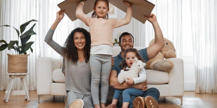 Pais e filhos brincando com caixa de papelão simulando casa em sala de estar.