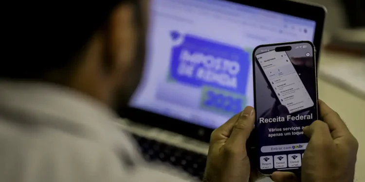 Homem acessa serviços da Receita Federal pelo celular enquanto visualiza portal do Imposto de Renda no notebook