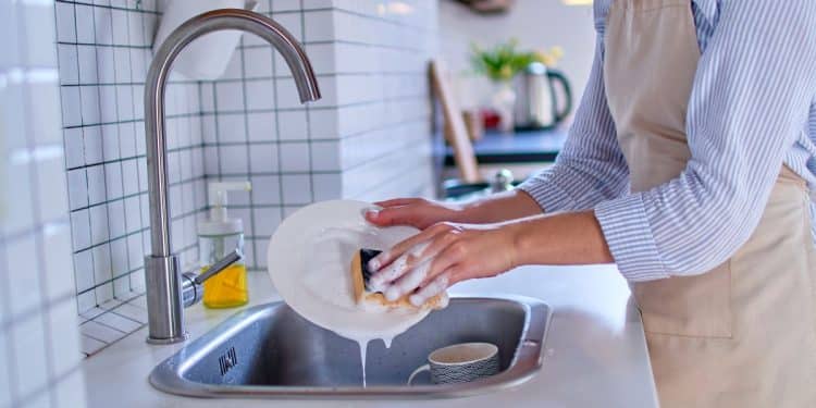 Pessoa lavando prato com esponja e detergente na pia da cozinha.