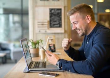 Homem de camisa azul comemora com punho cerrado olhando smartphone, notebook aberto em mesa de cafeteria