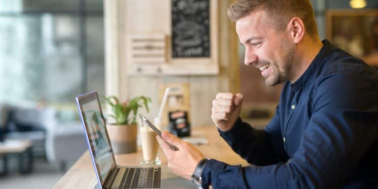 Homem de camisa azul comemora com punho cerrado olhando smartphone, notebook aberto em mesa de cafeteria