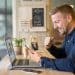 Homem de camisa azul comemora com punho cerrado olhando smartphone, notebook aberto em mesa de cafeteria