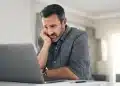 Homem preocupado olhando para o laptop em casa, lidando com o estresse, representando os desafios de carreira após os 40 anos.