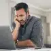 Homem preocupado olhando para o laptop em casa, lidando com o estresse, representando os desafios de carreira após os 40 anos.