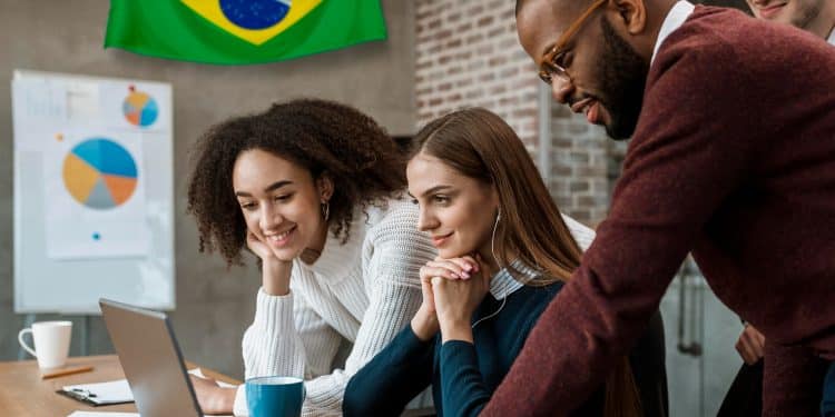Equipe diversificada de jovens profissionais reunida em torno de um notebook em ambiente corporativo com bandeira do Brasil ao fundo