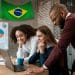 Equipe diversificada de jovens profissionais reunida em torno de um notebook em ambiente corporativo com bandeira do Brasil ao fundo