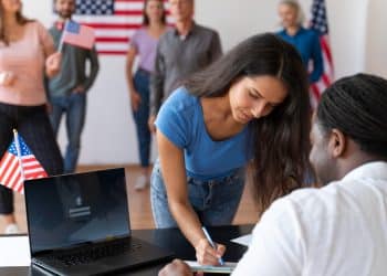 Empregos EUA sem faculdade - mulher assinando documento em processo seletivo com bandeiras dos Estados Unidos ao fundo