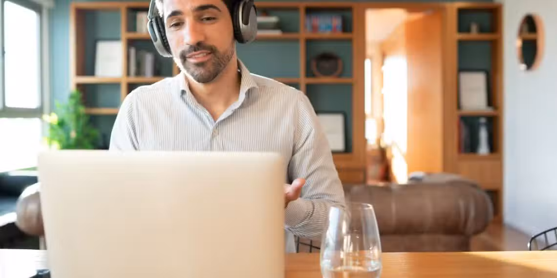 Homem de barba, camisa clara e fones de ouvido sorri durante videochamada em ambiente doméstico com estante ao fundo, representando o perfil do profissional brasileiro que atua de forma remota e valoriza a flexibilidade no trabalho