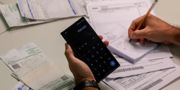 Consumidor fazendo contas em calculadora do celular cercado de faturas de energia elétrica com suspeita de empréstimo indevido