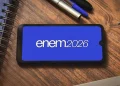 Tela de smartphone com o logo do Enem 2026 em fundo azul, com canetas e cadernos ao redor.