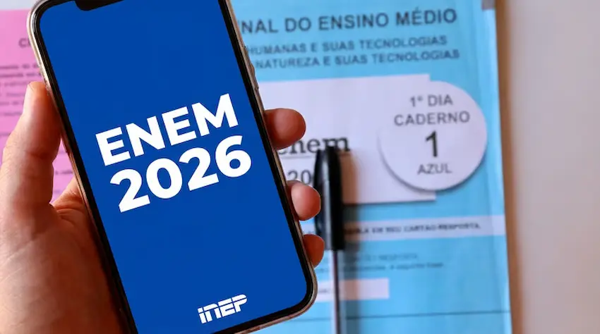 Pessoa segurando um celular com o logo do ENEM 2026 na tela, com a prova do ENEM 2026 ao fundo.