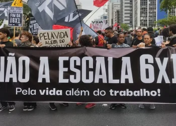 Manifestantes segurando faixa em protesto contra a escala 6x1, com bandeiras e cartazes sobre a vida além do trabalho.