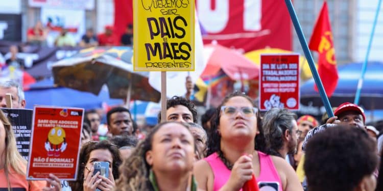 Multidão em ato sindical com cartaz amarelo "Queremos ser pais e mães presentes" e bandeiras da CUT