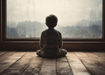 Silhueta de menino de costas, sentado no piso de madeira, observando janela grande com vidro embaçado pela chuva