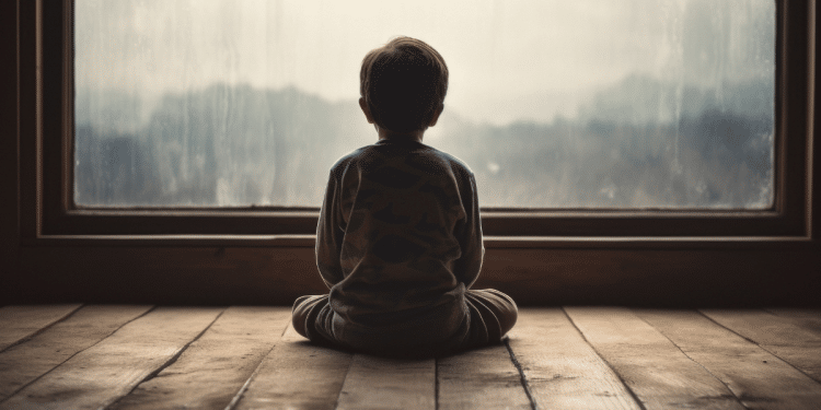 Silhueta de menino de costas, sentado no piso de madeira, observando janela grande com vidro embaçado pela chuva
