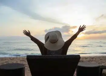Mulher de costas desfrutando da vista do mar ao pôr do sol, representando momentos de descanso e relaxamento em viagens durante o mês de maio de 2026.