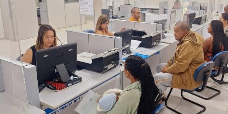 Servidores do INSS atendendo segurados em balcões com computadores em agência da Previdência Social, com placas indicativas e cadeiras para o público