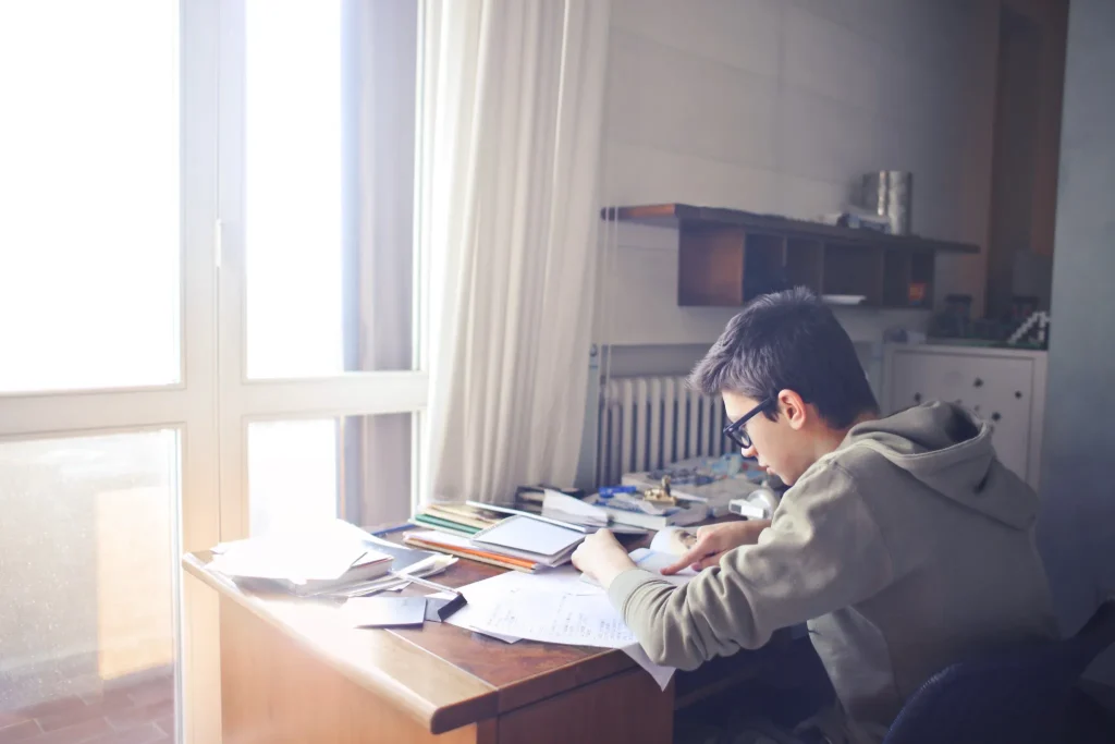 Jovem estudando em casa com cadernos e materiais sobre a mesa, representando aprendizado e atenção a regras da língua portuguesa.