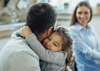 Pai de camiseta cinza abraça filha loira sorrindo com olhos fechados enquanto mãe observa ao fundo.
